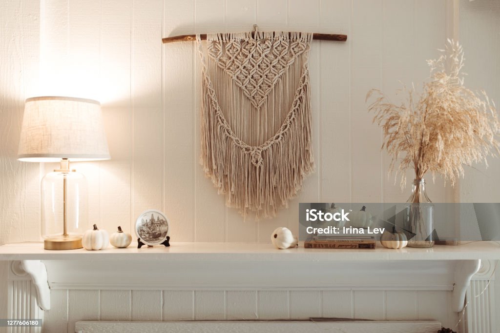 Thanksgiving home  decor - white wooden fireplace with wall macrame, cream and beige pumpkins, pine cones garland and rustic dry wheat bouquet. House fall decoration. Autumn Stock Photo