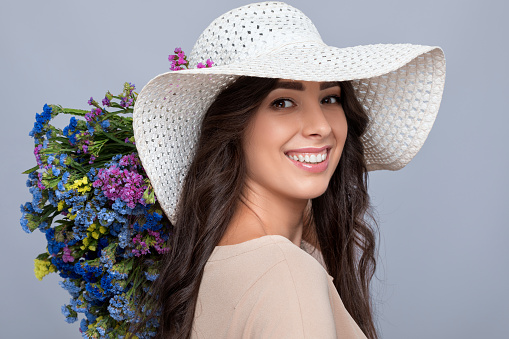 Portrait of a beautiful brunette girl with beautiful makeup and healthy clean skin. She holds a bouquet of colorful flowers. She's wearing a beige summer dress and hat. Makeup concept.