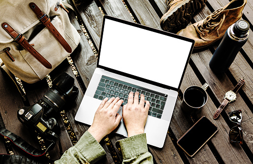 Traveler or travel photographer working on laptop computer outdoor among his equipment in the background (backpack, smartphone, boots). Captured from above (top view). Blank screen as copy space.