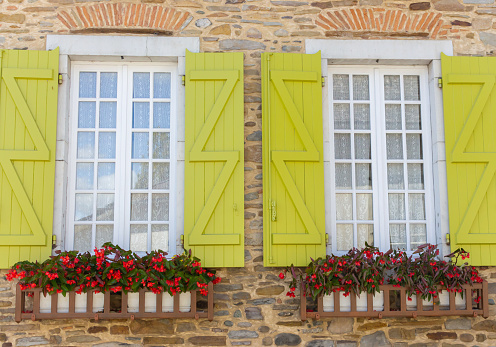 White wooden windows with flower in pots and light green shutters. Facade of ancient stone house in France. Typical exterior of medieval building, France. French vintage house. Old architecture concept.