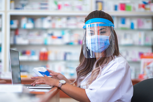 Young woman using laptop checking medicine and health products stock stock in hospital pharmacy or drugstore. Medical mask