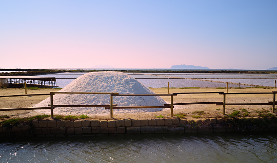 A hill of salt stands behind a canal and in front of salt pans and saltwater pools, at sunset near Marsala, Sicily