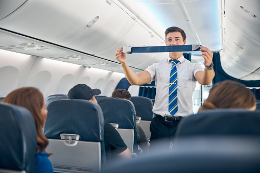 Close up portrait of young confident man flight attendant in business uniform demonstrating how fostering passenger seatbelt
