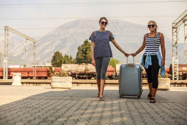 Low angle, front view of two young women standing at the train station, holding same piece of the luggage and smiling as a sign of excitement about the trip ahead of them.
