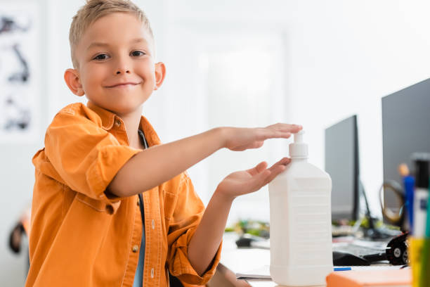 istock Selective focus of schoolboy using hand sanitizer and looking at camera in classroom 1277708350