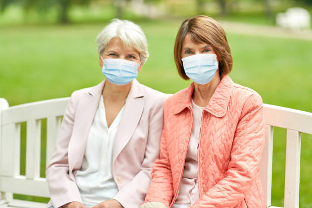 senior women in medical masks at park health, safety and pandemic concept - two senior women or friends in medical masks for protection from virus sitting on bench at summer park senior adult women park bench 70s stock pictures, royalty-free photos & images