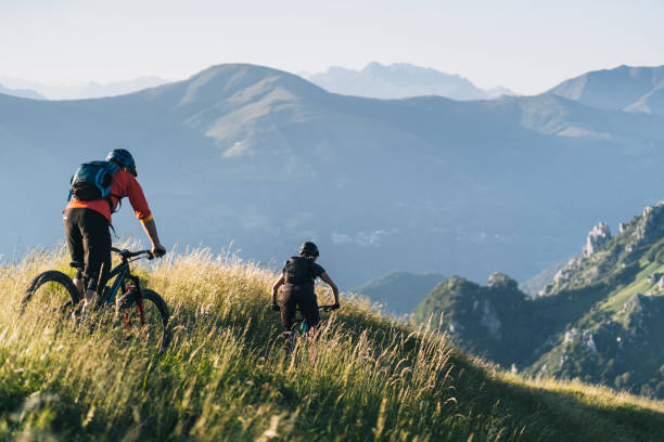 Mountain bikers ride down grassy mountain ridge Dolomites illuminated in the background mountain biking stock pictures, royalty-free photos & images