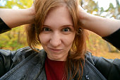 Funny portrait of a red-haired girl in the autumn forest. The beautiful girl put her hands on her head and bulged her eyes. Horizontal photography