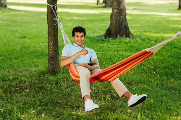 istock Smiling guy sitting on hammock in park and holds digital tablet 1277739495