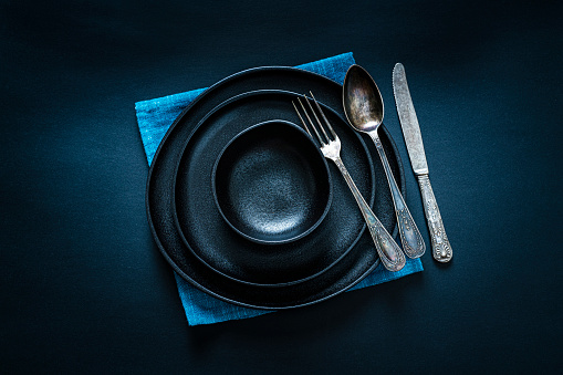 Overhead view of a black crockery on blue textile napkin shot on black background. A fork, spoon and table knife are beside the plate. Predominant colors are black and blue. Useful copy space available for text and/or logo on the plate. High resolution 42Mp studio digital capture taken with SONY A7rII and Zeiss Batis 40mm F2.0 CF lens