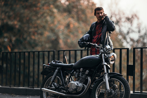 Caucasian male biker standing next to his motorbike, talking on his smart phone