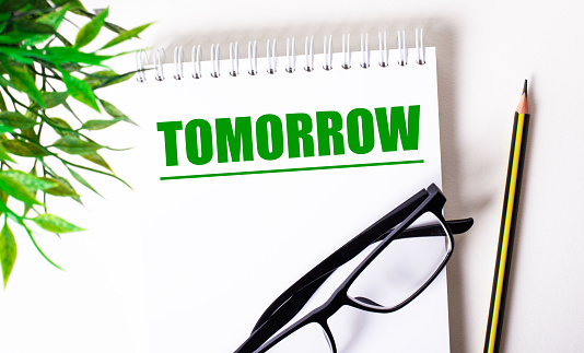The word TOMORROW is written in green on a white notebook next to a green plant, black-framed glasses and a pencil. Time concept