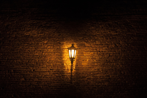 Vintage old street lantern lit with an orange light in front of a brick wall during a gloomy dark night.