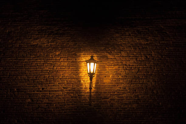 Vintage old street lantern lit with an orange light in front of a brick wall during a gloomy dark night. Picture of a street lamp in a street, at night, with a brick wall in background, projecting a yellow orange gloomy light. lanterns at night stock pictures, royalty-free photos & images