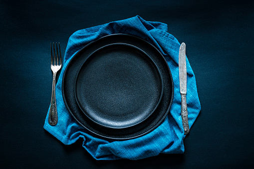 Overhead view of a black crockery on crumpled blue textile napkin shot on black background. A fork and table knife are at each side of the plate. Predominant colors are black and blue. Useful copy space available for text and/or logo on the plate. High resolution 42Mp studio digital capture taken with SONY A7rII and Zeiss Batis 40mm F2.0 CF lens
