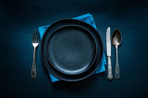 Overhead view of a black crockery on blue textile napkin shot on black background. A fork, spoon and table knife are at each side of the plate. Predominant colors are black and blue. Useful copy space available for text and/or logo on the plate. High resolution 42Mp studio digital capture taken with SONY A7rII and Zeiss Batis 40mm F2.0 CF lens