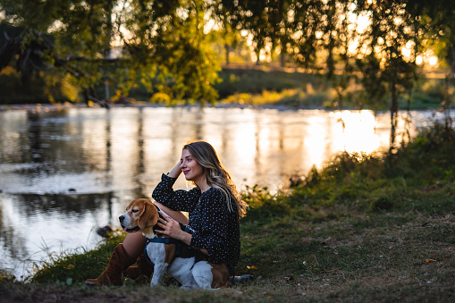 Beautiful young woman with her Beagle dog siting by the river during autumn sunset