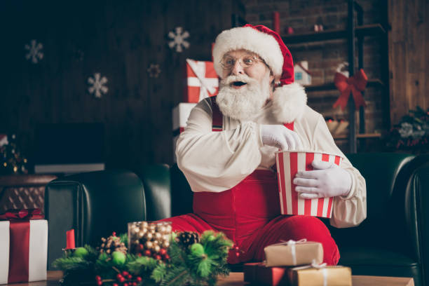 foto di eccitato barba grigia babbo natale sedersi divano guardare x-mas televisione di natale mangiare pop corn box stupito avvento indossare copricapo rosso in casa al chiuso noel ornamento - personal accessory cap christmas santa claus foto e immagini stock