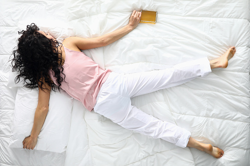 Top view of drunk female sleeping on white bedding holding alcohol bottle. Young woman resting after fun night party. Lady wearing light pants and pink shirt for sleep. Hungover and tired concept