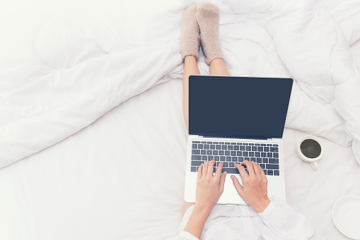 Close up legs women on white bed in the bedroom. Women working on laptop and drinking coffee in morning relax mood in winter season. Lifestyle Concept.