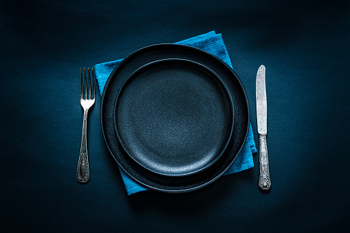 Overhead view of a black crockery on blue textile napkin shot on black background. A fork and table knife are at each side of the plate. Predominant colors are black and blue. Useful copy space available for text and/or logo on the plate. High resolution 42Mp studio digital capture taken with SONY A7rII and Zeiss Batis 40mm F2.0 CF lens