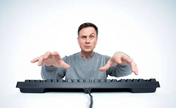 Man in a sweater is emotionally typing text on the keyboard, a look into the camera, on light background. Front view.