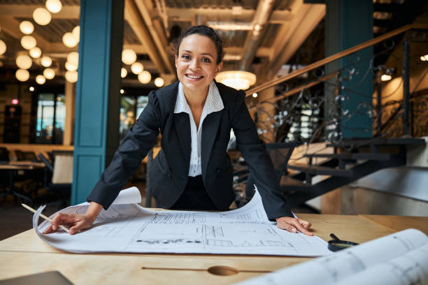 Positive delighted international female leaning on table Kind architect demonstrating her smile while working with the drawing of future office architect stock pictures, royalty-free photos & images