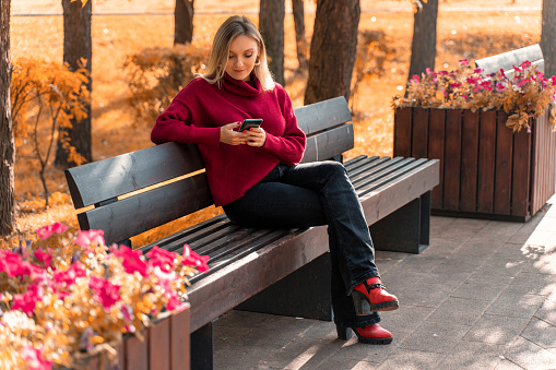 beautiful young blonde woman in a red sweater sits on a bench with a smartphone. autumn concept. internet in parks