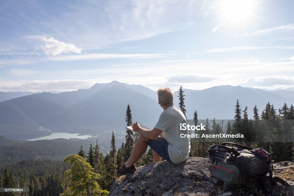 Male hiker relaxes on rock overlook, above valley He writes in journal. Forest and mountains in distance, valley Environmental Conservation Stock Photo