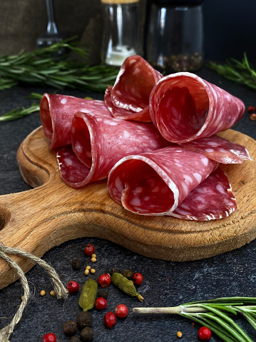 Fresh raw smoked salchichon sausage cut into thin slices on a wooden board with rosemary and pepper.