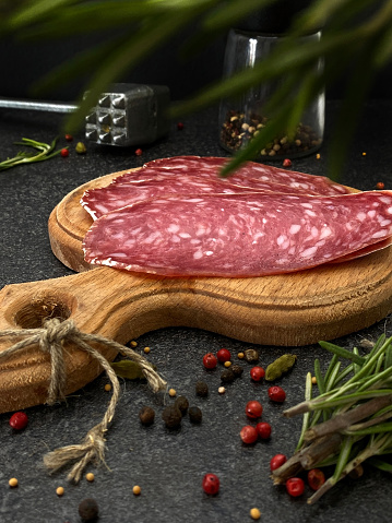 Fresh raw smoked salchichon sausage cut into thin slices on a wooden board with rosemary and pepper.