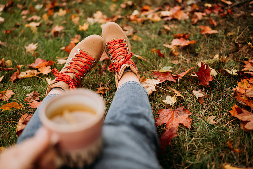 Woman drinking tea outdoors in autumn casual clothing close up of ceramic tea cup \nPhoto taken outdoors in nature in sunlight. WOMan wearing autumn winter boots