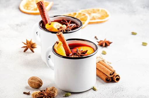 Mulled wine in white metal mugs with cinnamon, spices and orange on gray background, traditional drink on winter holiday. Copy space