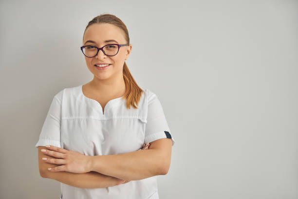 Cheerful female cosmetologist standing against white background Charming young woman beautician in glasses looking at camera and smiling while crossing arms over her chest aesthetician photos stock pictures, royalty-free photos & images