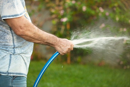 man watering flowers and grass with hose in the garden on a hot summer day.