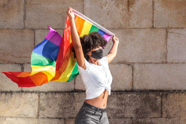 Woman with face mask and afro hair holds up the lgbtq flag of gay pride that moves with the wind stock photo