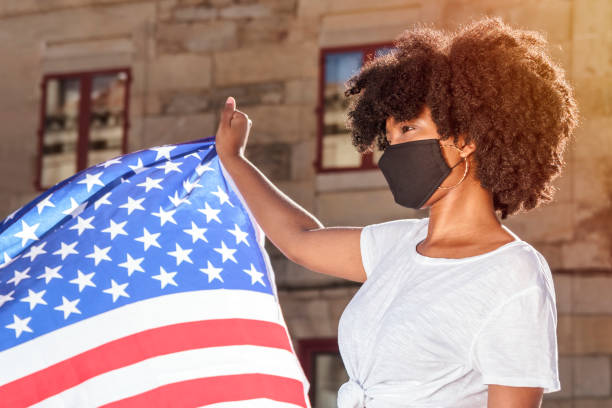 Black woman with mask and afro hair holds the American flag under the sunlight stock photo