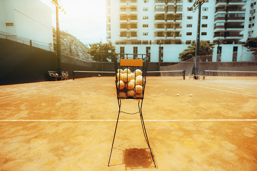 View of an empty outdoor tennis court with a selective focus on a basket on an orange clay ground, in the foreground, full of yellow balls for training; warm sunny afternoon