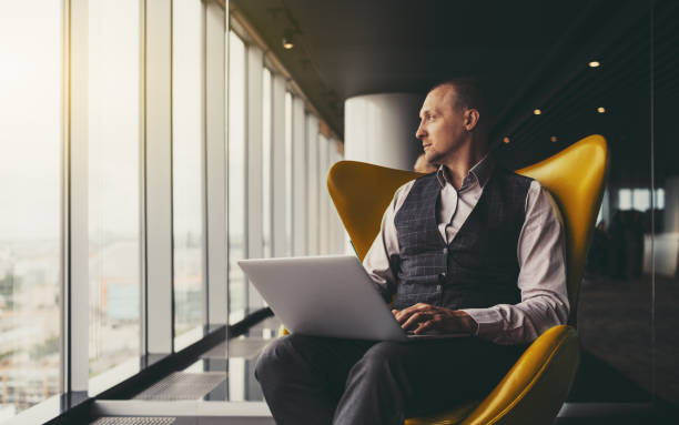 Businessman on armchair with laptop A confident relaxed successful mature man entrepreneur sitting with a laptop on an orange armchair and thoughtfully looking aside on an urban skyline outside the window; a copy space place on the left rich man stock pictures, royalty-free photos & images