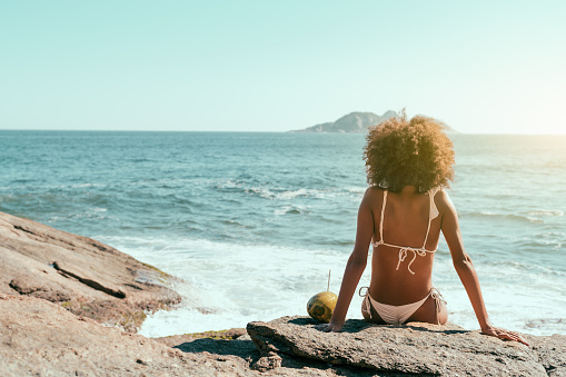 View from behind of a young African-American female with curly hair, in a swimsuit sitting on a rock near the beach of a resort and enjoying the sun, observing seascape, relaxing; a coconut near her