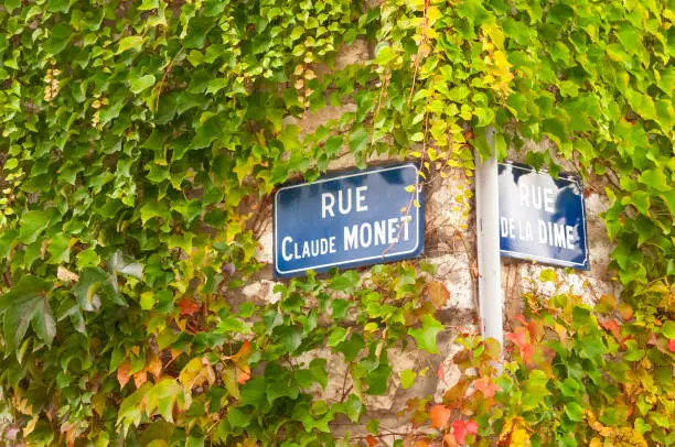 Photo of Close up of street signs on ivy covered walls at the corner of Rue Claude Monet (named after the famous impressionist painter who lived in the town) and Rue De La Dime in Giverny, France.