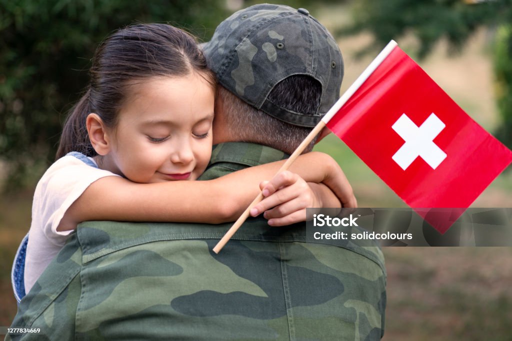 Swiss Soldier And His Daughter Little girl hugs the soldier father and holding Swiss flag. Swiss Military Stock Photo