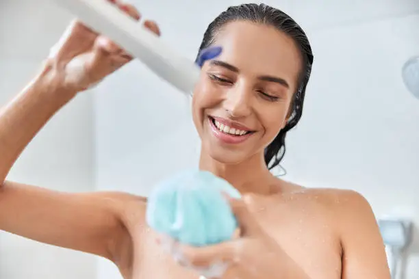 Beautiful lady squeezing shower cream on exfoliating sponge and smiling while taking shower at home