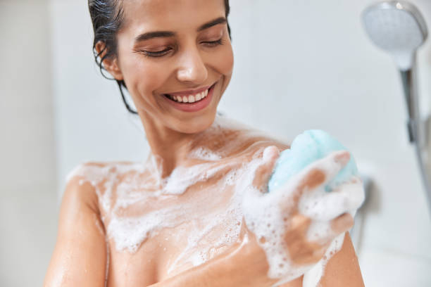 Cheerful young woman using exfoliating loofah while taking shower Beautiful lady with foam on her skin washing body with bath sponge and smiling cleaning sponge photos stock pictures, royalty-free photos & images