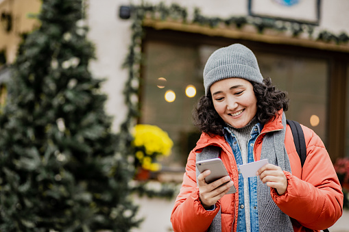 Photo of young woman standing on the street during winter, smiling and holding smart phone and credit card