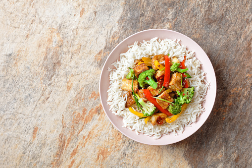 Japanese dish katsu chicken curry with vegetables: broccoli, red and yellow sweet pepper and parsley over jasmine rice served on a pink plate on a stone background served with chopsticks, close-up