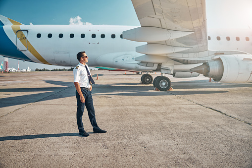 Joyful young man in uniform is relaxing in sun on runway near passenger plane before boarding
