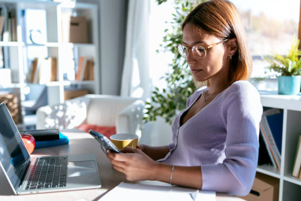istock Beautiful young business woman using her mobile phone while working with computer in the office at home. 1277892368