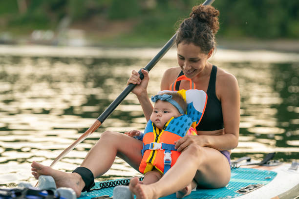 Adorable Baby Girl on Paddle Board with Her Mom An athletic Pacific Islander woman is paddle boarding with her baby daughter sitting between her legs. The child is wearing a lifejacket. They are enjoying a relaxing summer evening at the river. The woman is looking down and smiling at the adorable child. river swimming women water stock pictures, royalty-free photos & images