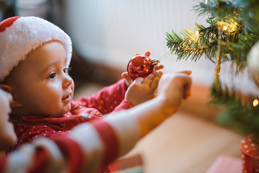 Portrait of a beautiful little baby girl that is wearing a Santa Claus hat and playing with the Christmas tree decoration